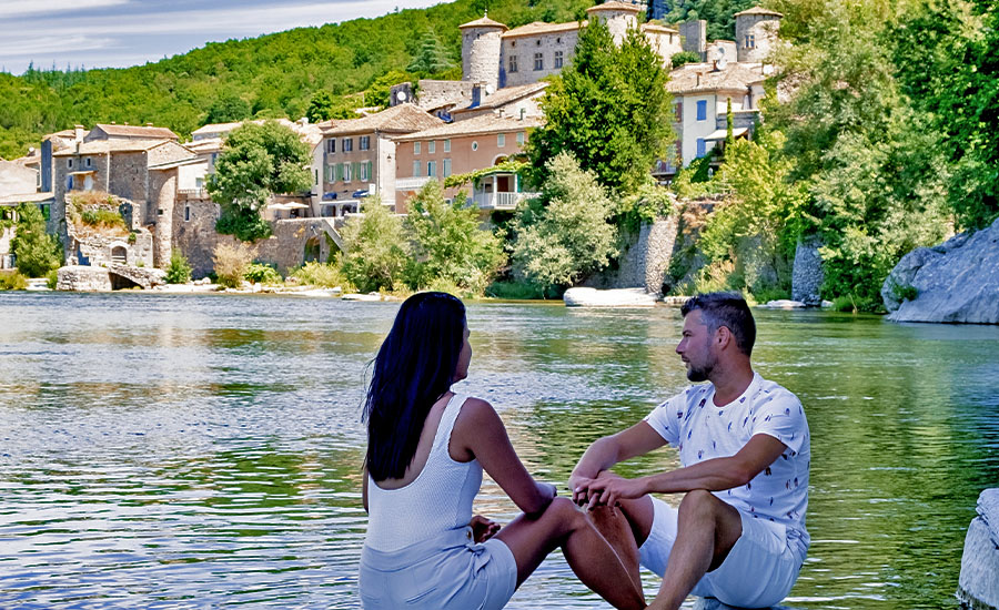 Couple assis au bord de la rivière face au village de Balazuc en Ardèche lors d’un séjour nature